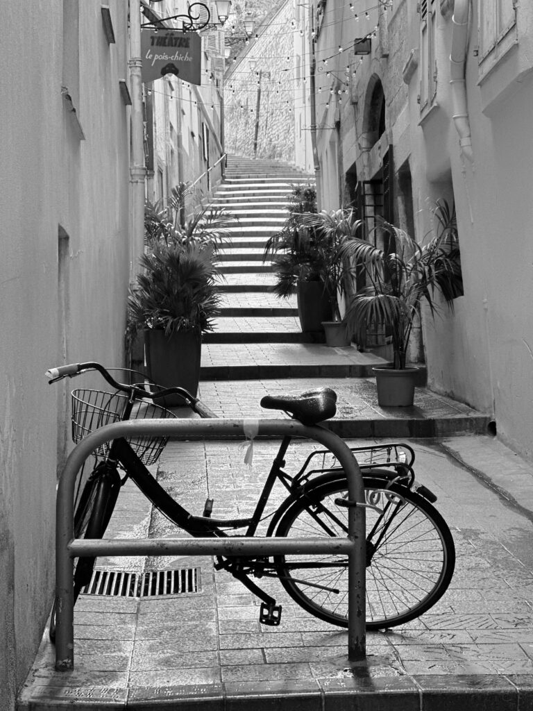 "Black and white photograph of a narrow cobblestone alleyway in Old Town, Nice, France. A vintage bicycle is parked against a metal railing in the foreground. Stone steps lead up between tall buildings, adorned with potted plants and hanging lights. A sign reading 'Théâtre le pois-chiche' hangs above, adding to the historic and charming ambiance of the scene."